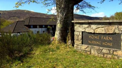 Outside a building in the background, with a tree and wall in the foreground. There is a sign on the wall that reads 'Home Farm Nursing Home'.