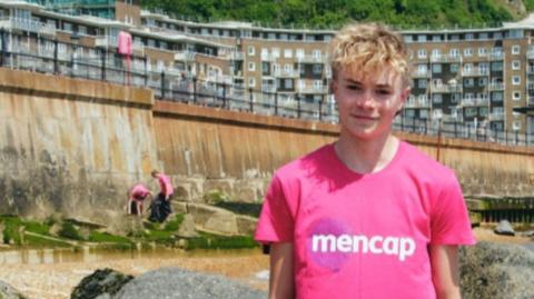 A young boy with blonde hair is wearing a pink t-shirt with white letters bearing the word Mencap on it. In the background is a tall sea wall and in the distance is a tower block with several floors.