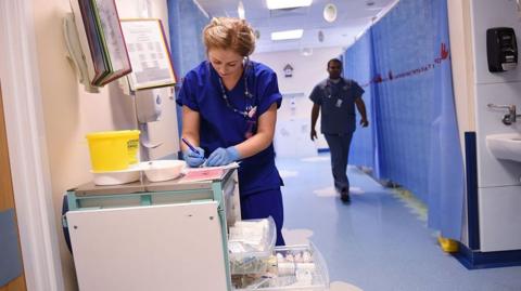 Member of staff filling in notes in hospital ward corridor in blue overalls and other doctor walking in background along curtained bays