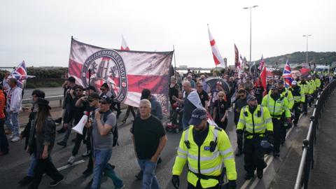 A group of protesters carrying a flag which reads English Defence League, marching along a road. They are marshalled by police officers in hi-vis jackets. 