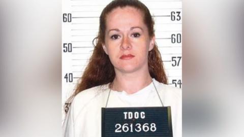 A woman with red hair stands in front of a sign that shows a person's hight, she wears a sign around her neck reading "TDOC 261368"