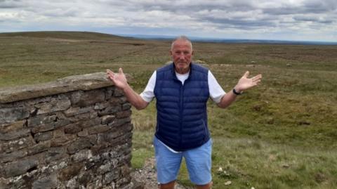 Lord Ian Botham stands outdoors on open moorland, with rolling hills, dry-stone walls and countryside stretching into the distance behind him.