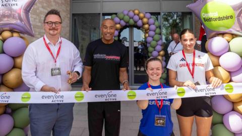 Four people stand behind the white ribbon which is branded with "everyone active" and "Central Bedfordshire Council." They are: Steve Watkins, Colin Jackson, Maisie Summers-Newton and Olivia Newman-Baronius. The councillor holds the scissors open. On either side of them, and across the entrance doors, are pastel coloured balloon arches.