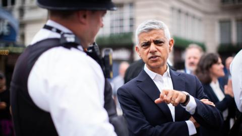 London Mayor Sadiq Khan, wearing a dark suit and white shirt, gestures while speaking to a uniformed police officer in the street, with several people blurred in the background.