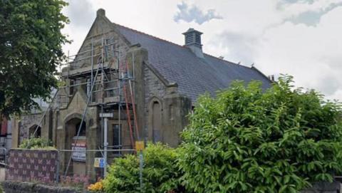 A church with scaffolding on the exterior. It is set back from the street and surrounded by a low, stone wall and shrubbery.