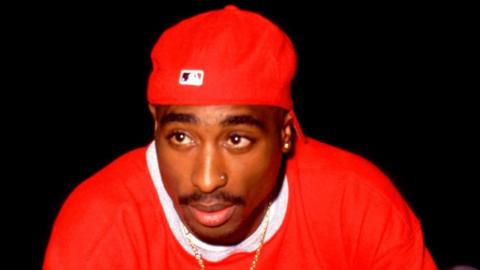 Tupac Shakur is seen wearing all red, including a red ball cap, and a gold chain. 