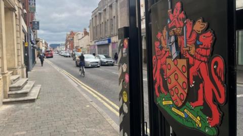 Lower Eastgate Street with cars and a cyclist going along the road and a gate in the foreground with a red, medieval crest