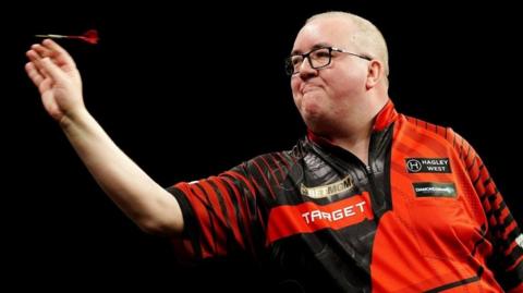 Stephen Bunting is throwing a dart. A bald man with glasses, he wears a red and black patterned collared shirt adorned with various sponsors' logos.