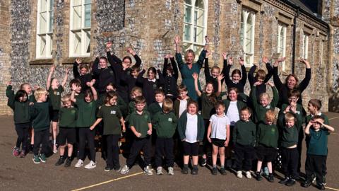 Two rows of school children from Antingham and Southrepps Primary School standing outside their school on the playground. They are all wearing school uniform which consists of dark green jumpers and cardigans, some with white T-shirts. Most are smiling and looking at the camera with their arms in the air.