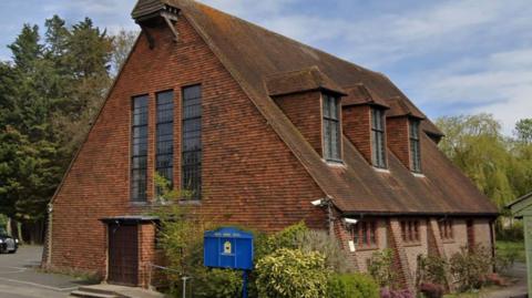 Surrey Muslim Centre. It is a brick building which has a pitched roof with three dormer windows. 
At the front three long windows are above the door. 
Shrubs are growing at the side