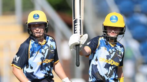 Two female cricketers are wearing yellow helmets and blue and black tops while the player on the right holds up her bat