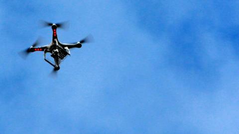 A drone camera with three red anti-collision lights flying in the air. The sky is blue with no clouds.