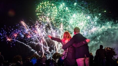 A man and a woman are standing with their arms around each other facing a fireworks display.