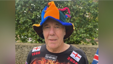 A head and shoulder image of Barry Illing from Gloucestershire wearing a joker hat and a black t-shirt with England flags on and red writing stating 1966 ad 2026, with a green bush in the background. 