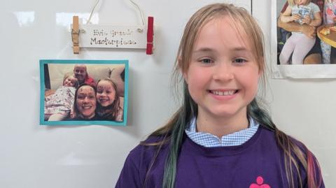 A young girl with long blonde hair stood in front of two family photographs.  She is wearing a purple top with the logo of the bereavement charity, Stand By Me