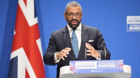 Sir James stands at a podium in front a deep blue background  -  a union jack flag is prominent on the left of the image. He is wearing a dark blue suit, a patterned blue and green tie and a white shirt. 