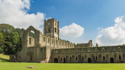 A large medieval stone abbey stands in the centre of a wide, bright green lawn.  The abbey is in ruins, with tall walls, large pointed Gothic arches, and rows of narrow windows. Behind the ruins are tall green trees, while above them is a bright blue sky with large white clouds.
