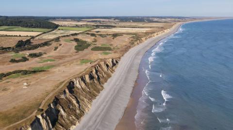 Birds-eye view of Norfolk coastline with cliffs, sea and horizon.
The cliffs are sandy-coloured and areas of trees and grassland are dotted on its top. Many of the fields are parched dry.