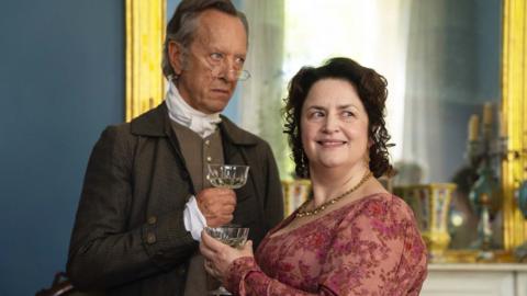 Actors Richard E Grant and Ruth Jones as Mr and Mrs Bennet, dressed in Victorian costumes and holding wine glasses, standing in front of a large golden framed mirror.