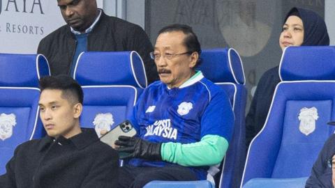 Vincent Tan wears a blue football shirt while sat in a blue seat inside stadium. He holds a mobile phone in hand.