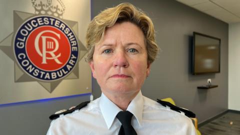 A police chief constable, a woman with blonde hair just past her ears, has a stern look on her face as she sits in a room in a police station. She is in uniform - a white shirt with a black tie and black detailing on the shoulders - and the Gloucestershire Constabulary logo is behind her.