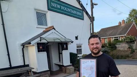 A man called Lee Wheaton stands outside a pub called the New Fountain Inn, where he is landlord. He is wearing a black T-shirt and is holding a framed certificate. He has a dark beard and short black hair.