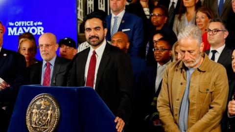 A man stands at a podium in a suit, another man holds his head down standing next to him. 