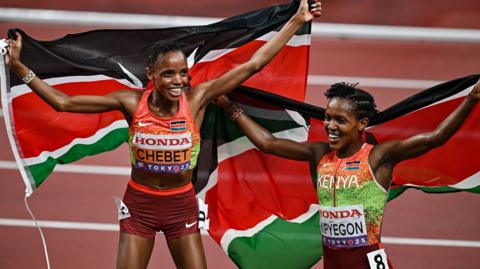 Beatrice Chebet and Faith Kipyegon, wearing orange, green and maroon Kenyan running kit, hold Kenyan flags behind them as they celebrate on a terracotta-coloured running track