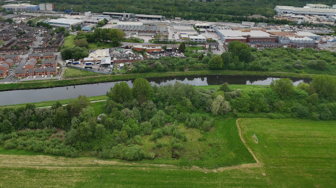 River with green fields on either side and dense trees. There is housing and commercial or industrial buildings in the background. The view is elevated.