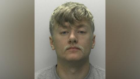 A police mugshot of a 23-year-old man with blonde hair and a moustache. He is wearing a grey T-shirt and is standing against a painted white background.