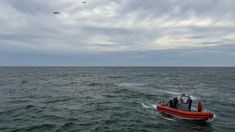 A bright red coast guard vessel at sea with crew onboard, and two aircraft in the distant sky overhead