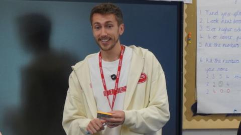 A man in a white hoodie and shirt standing in front of a classroom. There is a blackboard behind him. He is holding a pen and some sticky notes.