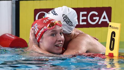 Keanna MacInnes, in a red swim cap, is hugged by an opponent while still in a swimming pool at the conclusion of a race