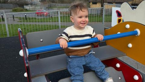 Photograph of baby Noah on a ship in a playground. He wears blue jeans and a navy, cream and tan striped long sleeve t-shirt. He is two years old and has brown short hair and blue eyes. He holds on to the rail and smiles. 