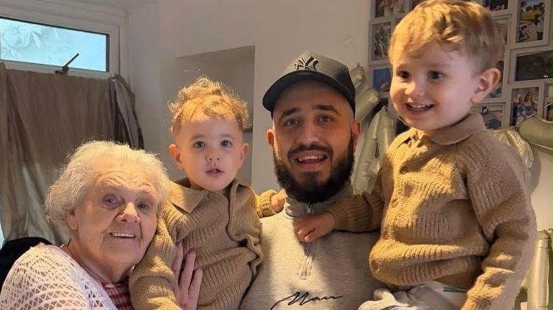 Tarik Al Roshoq (centre) holds both of his sons in his arms. He wears a cap and a grey sweatshirt. The boys were tan jumpers and smile at the camera. The grandmother (left) leans into the three. She has silver short hair and blue eyes. 