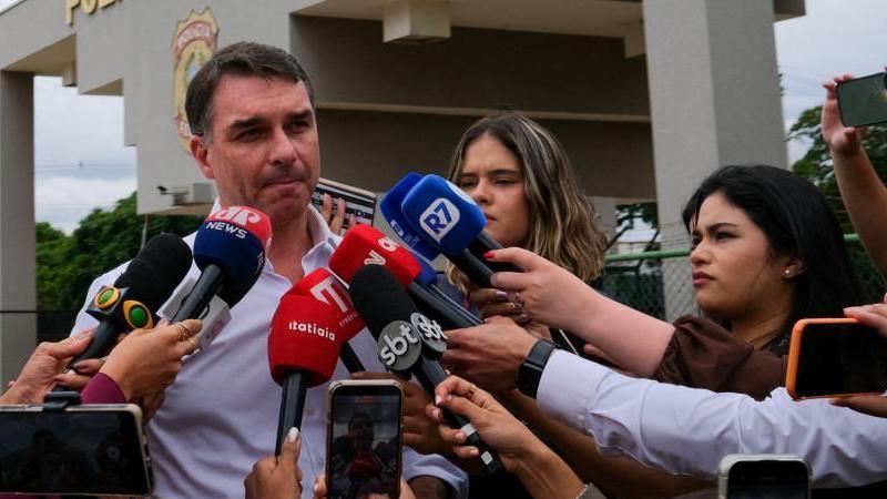A file photo from 16 December shows Senator Flávio Bolsonaro speaking to the journalists after visiting his father at a Federal Police building in Brasilia.
He is wearing a light-blue shirt and in the background the police building can be seen. Journalists are proffering their microphones at Flávio Bolsonaro.
