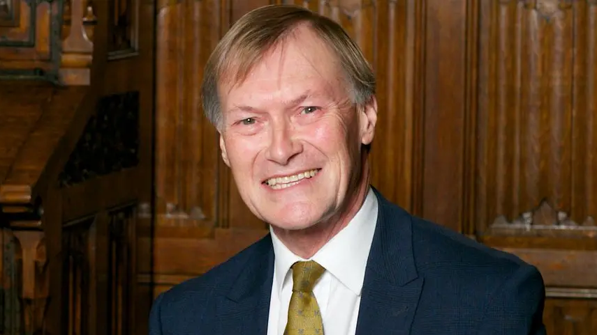 Sir David Amess, smiling, wearing a navy suit and an olive green tie in front of wooden panels.