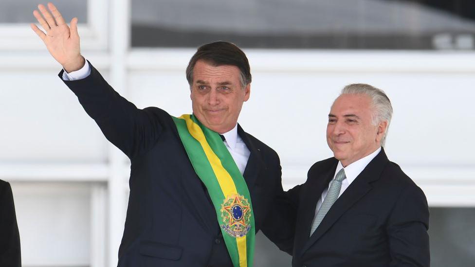 Jair Bolsonaro, wearing the presidential sash over a black suit, waves to supporters during his swearing-in ceremony. Next to him is the outgoing Brazilian president, Michel Temer