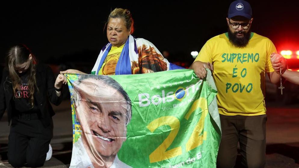 Supporters of former Brazilian President Jair Bolsonaro kneel and hold hands outside his home, awaiting the Supreme Court's final deliberations in the trial where he faces charges of plotting a coup following his electoral defeat, in Brasilia. They are holing a banner with a photo of Bolsonaro. A beaded man is kneeling. His T-shirt reads: "the people are supreme".