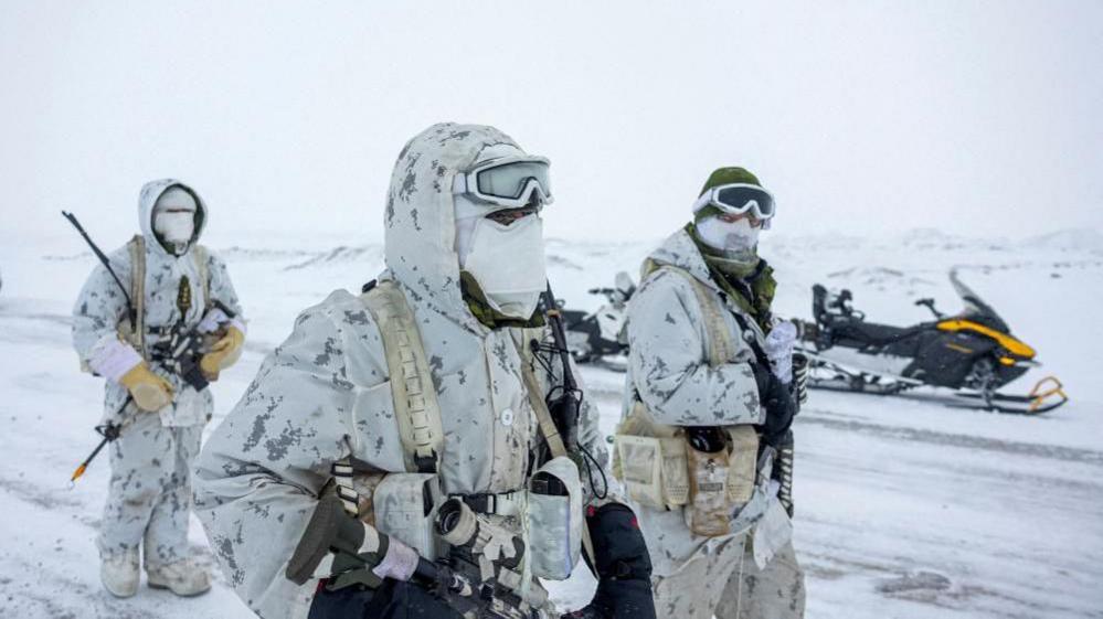 An image showing three Canadian military members in a snowy landscape, wearing white camouflage uniforms, face covers, gloves and holding various types of ammunition as they walk towards the right of the frame. Behind them is a parked snowmobile. 
