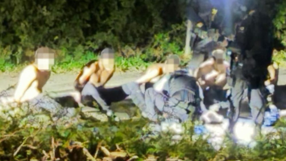 A photo of suspects with hands behind their backs on the ground, surrounded by police.
