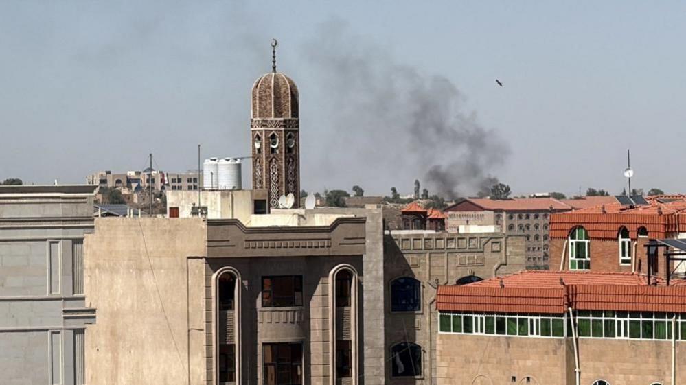 Smoke rises from buildings in a Yemeni city after reported strikes, with a towered structure and densely packed rooftops visible under a clear sky