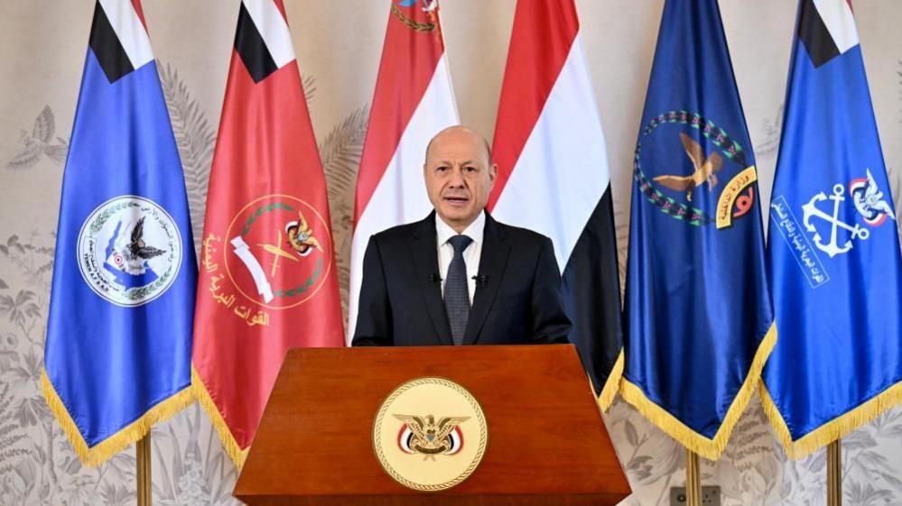 Yemeni President Rashad al-Alimi delivers a televised address from a podium, flanked by Yemeni and military flags