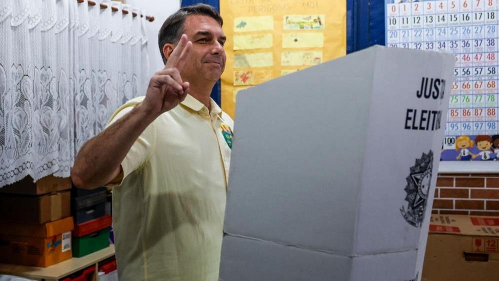 Flávio Bolsonaro raises two fingers while standing beside a voting booth marked “Justiça Eleitoral” after casting a ballot at a polling station in Rio de Janeiro
