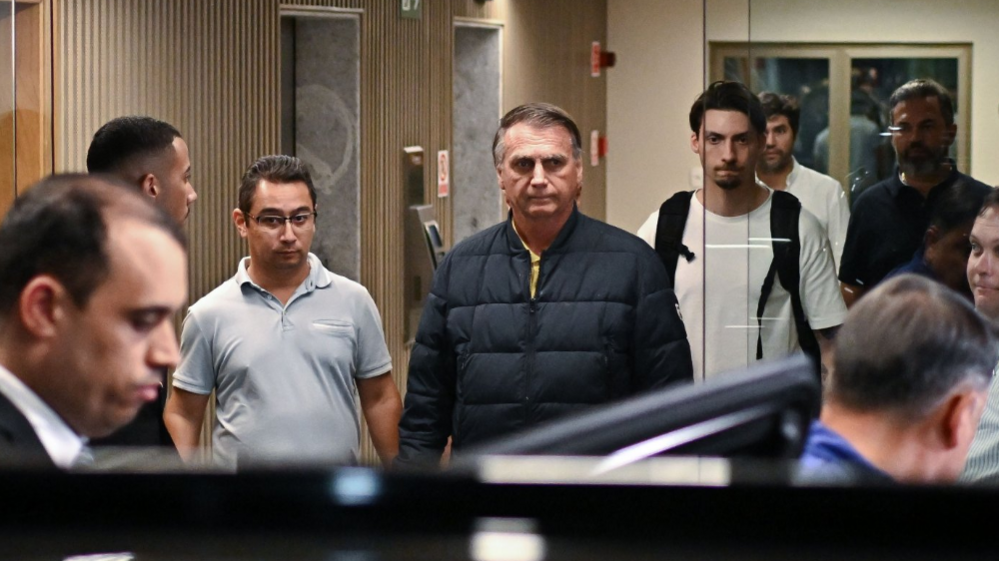 Former Brazilian President Jair Bolsonaro is wearing a navy blue parka as he leaves a building with several people around him. He is seen through glass doors leaving a lobby with elevators behind him. He has a grim expression.