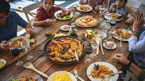 A family sits around a table enjoying a large pan of seafood paella (Credit: Getty Images)