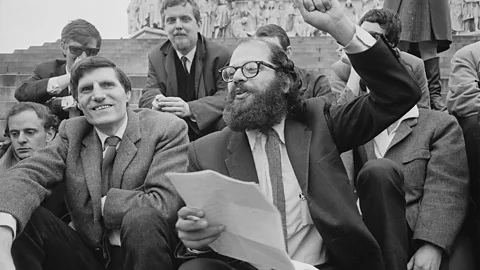Getty Images Black and white image of Allan Ginsberg, reading sheets of paper with his other fist raised, sat alongside fellow Beat poets (Credit: Getty Images)