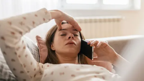 Woman lying back on a sofa with one hand on her forehead and the other hand holding a mobile phone to her ear