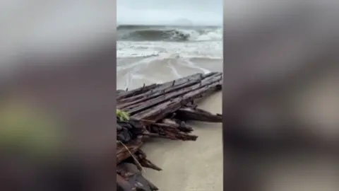 A wooden shipwreck washed up on a beach with big waves in the background.