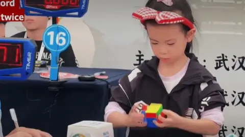 A screen shot of a young girl competing in a Rubik's cube competition with a timer in the background